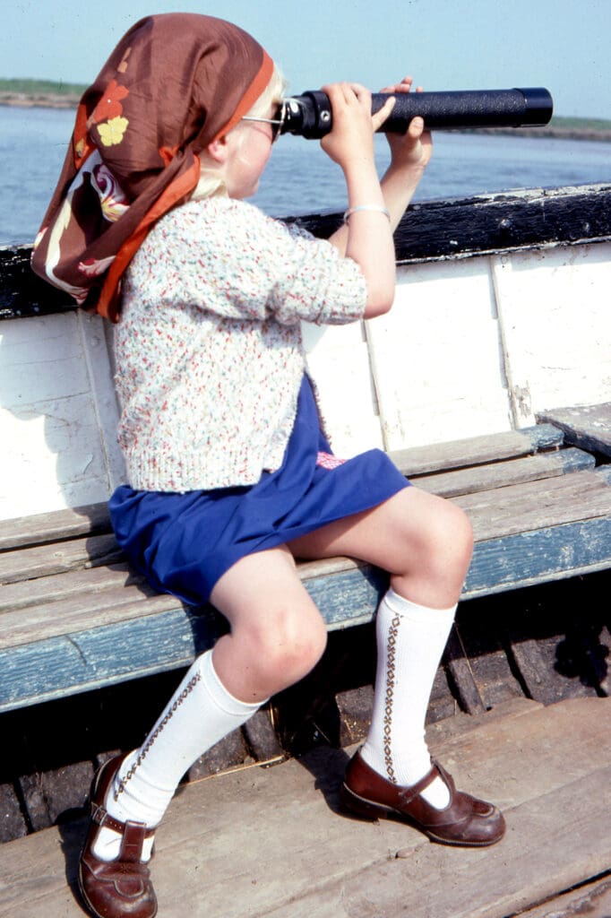 A young girl in a brown headscarf is sitting on a boat, facing away from the camera and looking through a hand-held telescope.