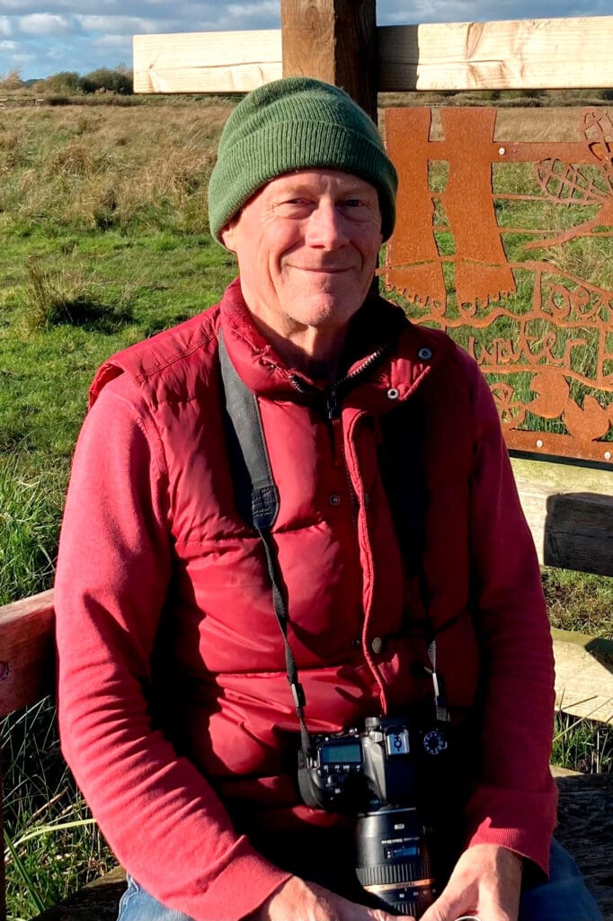 A man in a green hat and red coat, with a camera on a strap around his neck, sitting on a wooden bench with an ornate back, smiling at the camera.