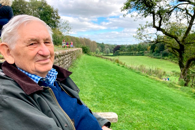 A man sitting on a bench in a green landscape on a sunny day.