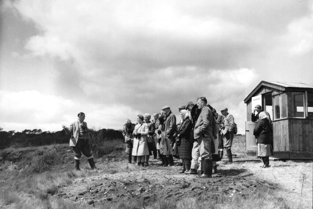 A black and white photo showing a group of people standing next to a wooden bird hide.