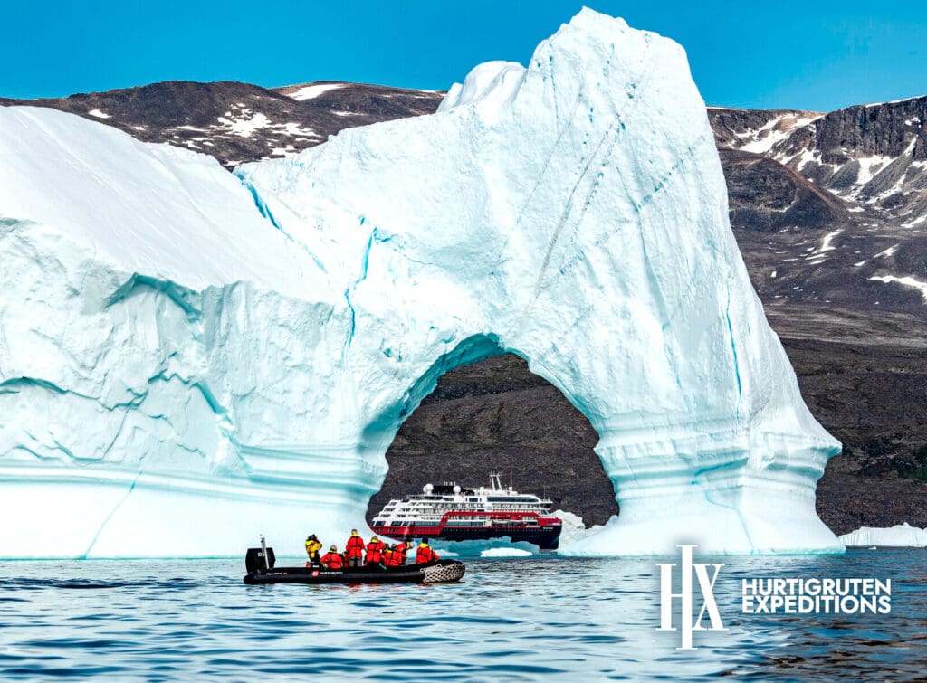 Boats passing an archway of ice rising from the sea, mountains in the background