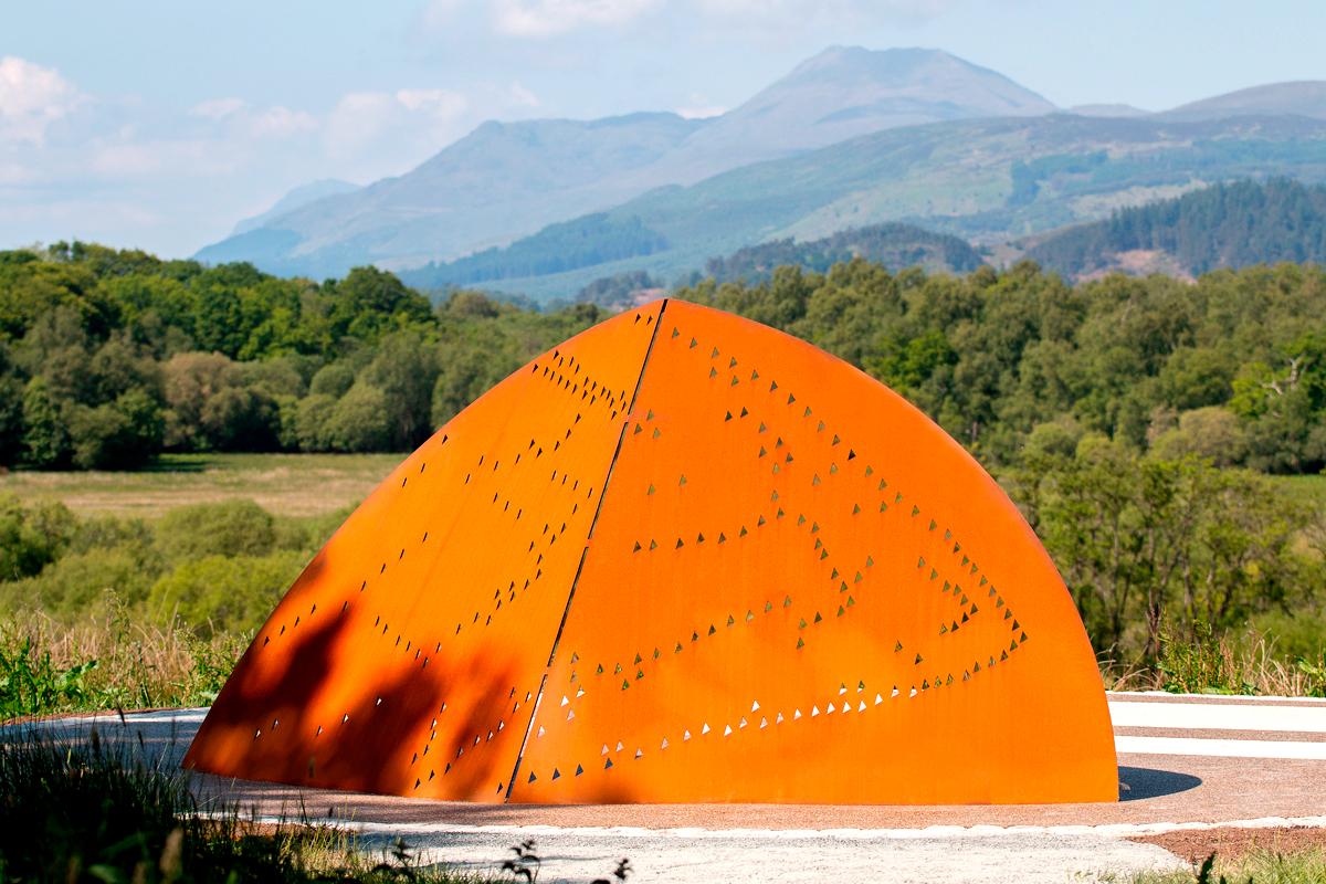 An orange dome-shaped sculpture with the pattern of a skein of geese cut into it, forest and mountains in the background.