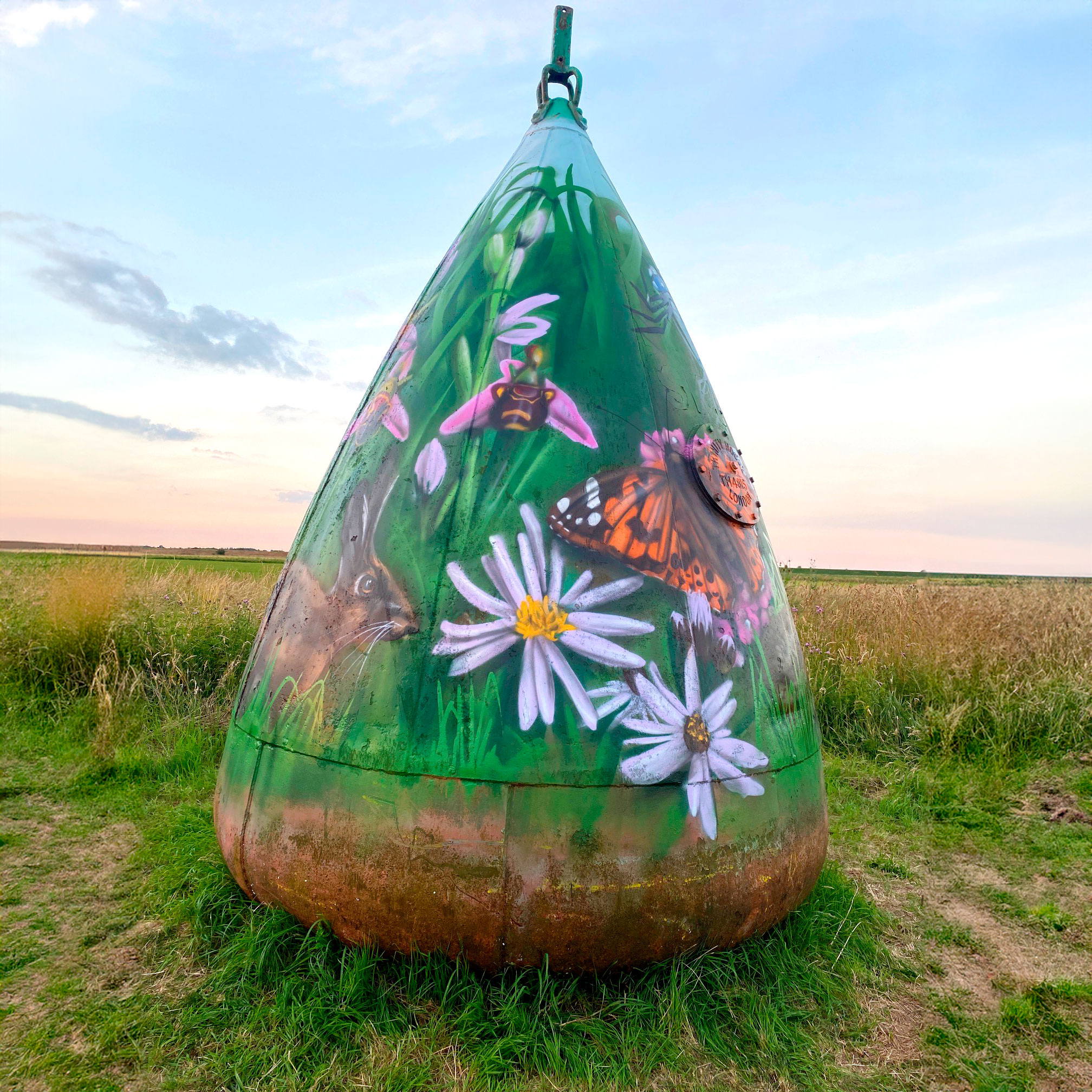 A shipping buoy which has been painted with images of plants and animals, on grass surrounded by reedbeds.