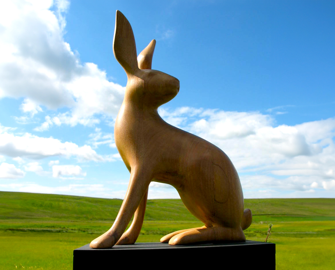 A wooden sculpture of a rabbit, green fields and blue sky in the background.