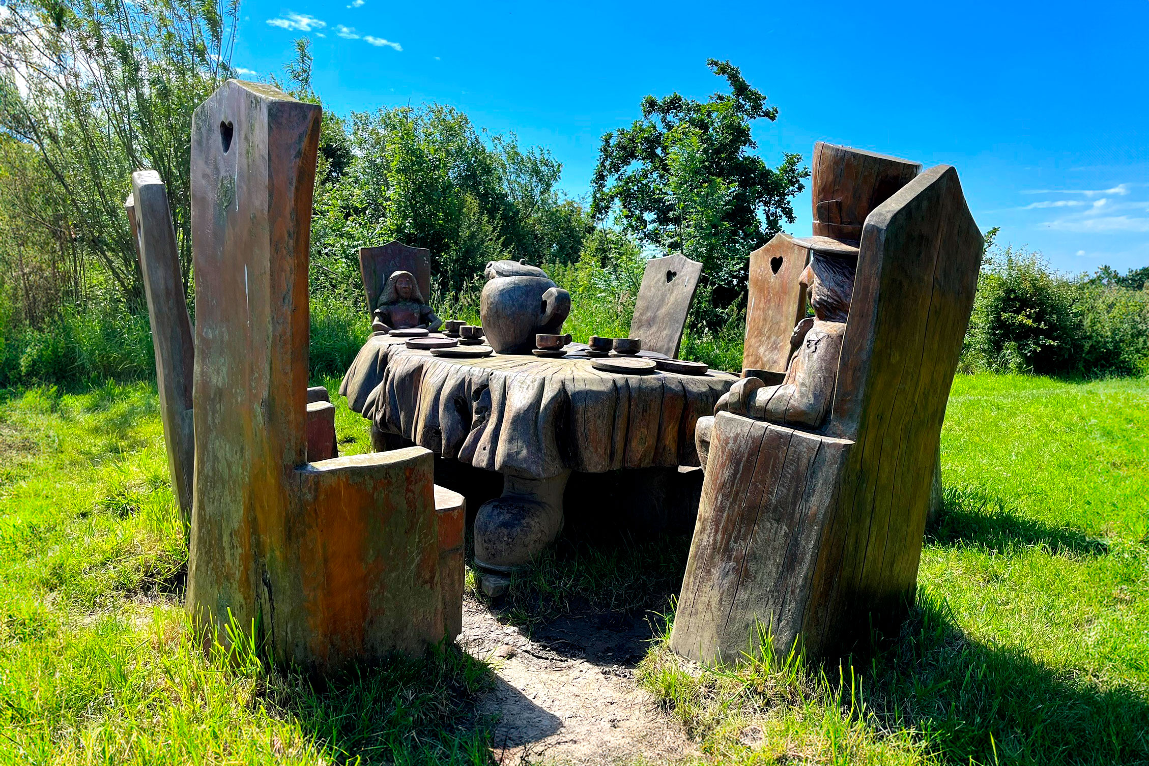 Wooden sculptures representing the Mad Hatter's Tea Party from the book Alice in Wonderland, set on a grassy field with trees in the background.