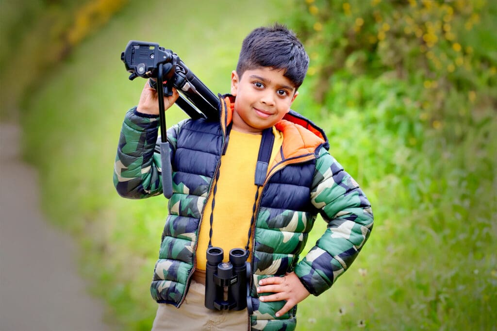 Aneeshwar stands in front of a hill with a tripod on his shoulder and binoculars around his neck.