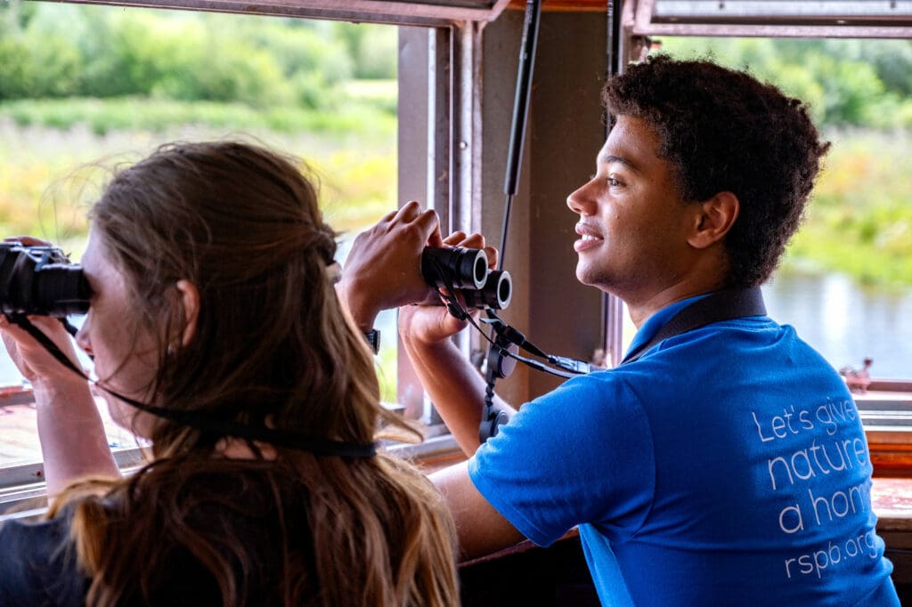 Two young people holding binoculars and looking out of the window of a wooden birdwatching hide