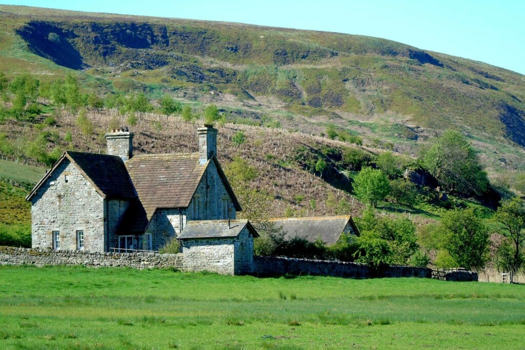 an old stone building with sloping roofs and chimneys in a green valley, hill and trees in the background.