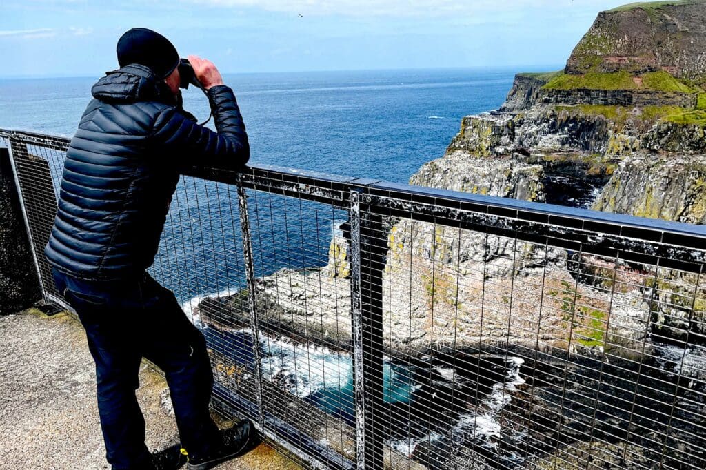 A man wearing a blue coat is leaning on a railing, looking across at a cliff through a pair of binoculars