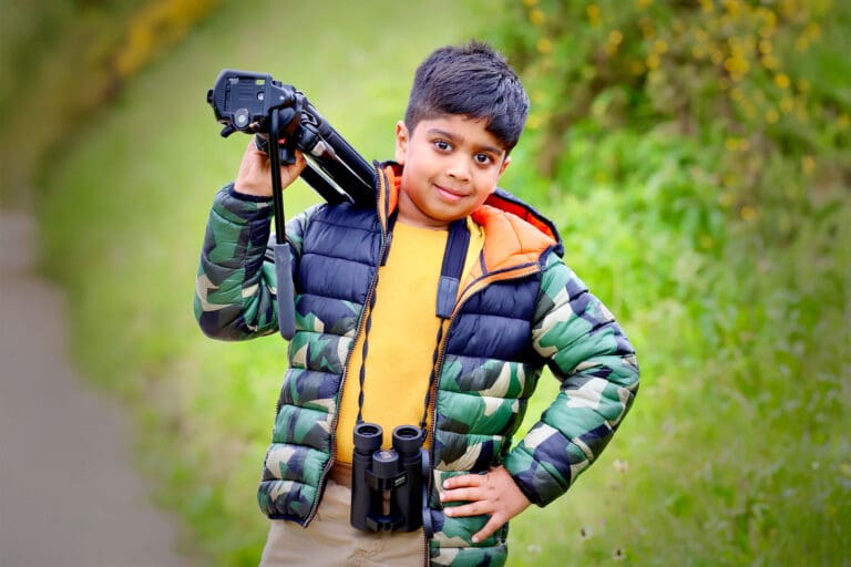 A boy poses with binoculars around his neck, while balancing a camera tripod on his shoulder with one hand