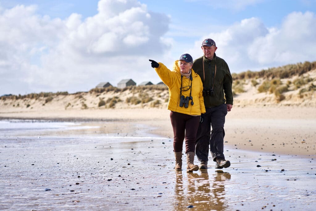 Jacinta is pointing at something in the distance as she and Dave walk along the shore