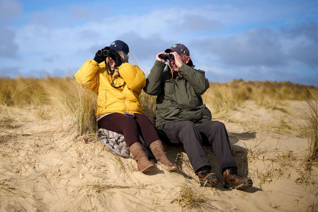 Jacinta and Dave are sitting on the dunes, both looking through their binoculars