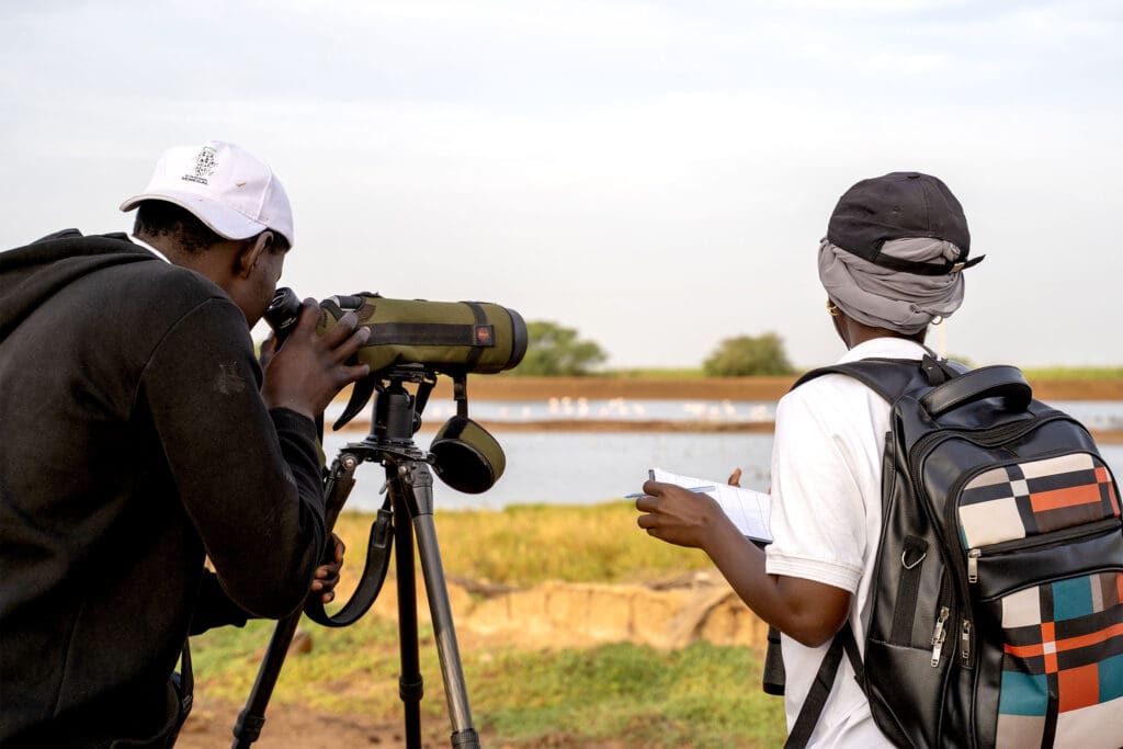 A man looks through a telescope at waders in the water. A woman next to him is writing notes.