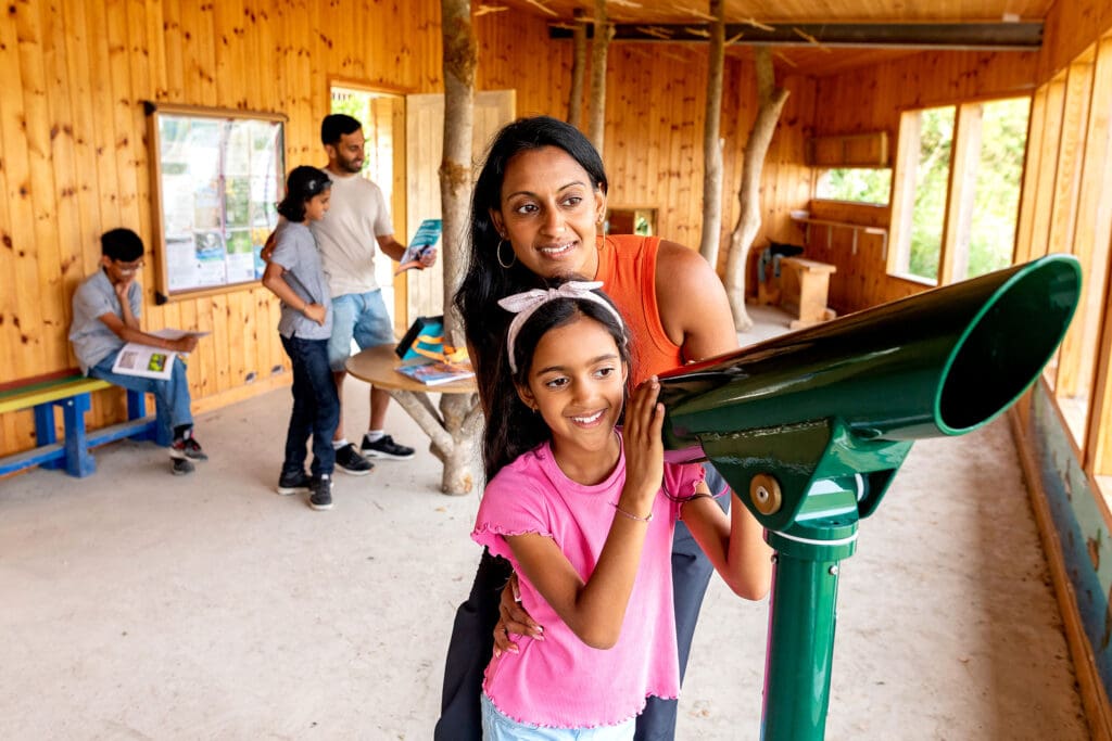 A woman and young girl stand in a bird hide next to a telescope, looking outwards