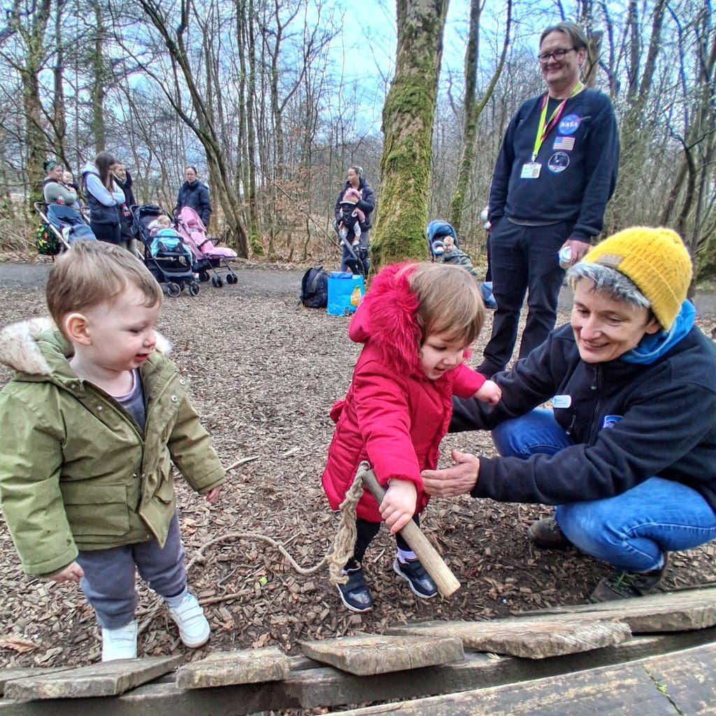 Carers and young children in a wooded area, playing with a large wooden musical instrument