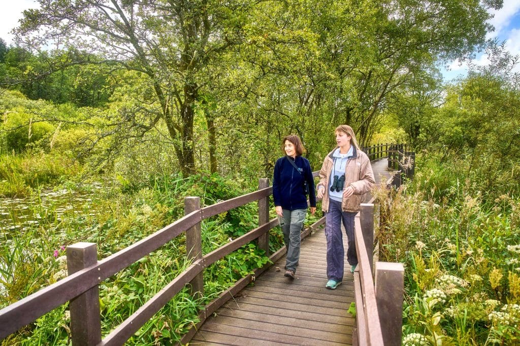 Two women walk a boardwalk surrounded by trees and greenery