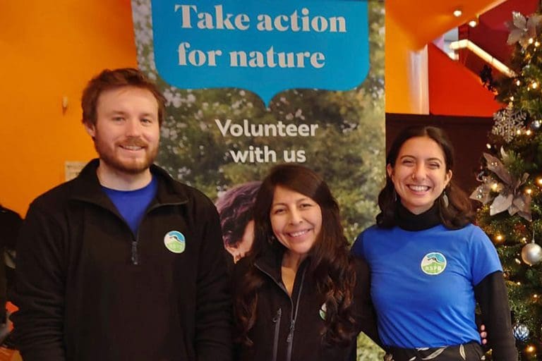 Three volunteers in RSPB t-shirts and fleeces stood in front of a 'Take action for nature' sign