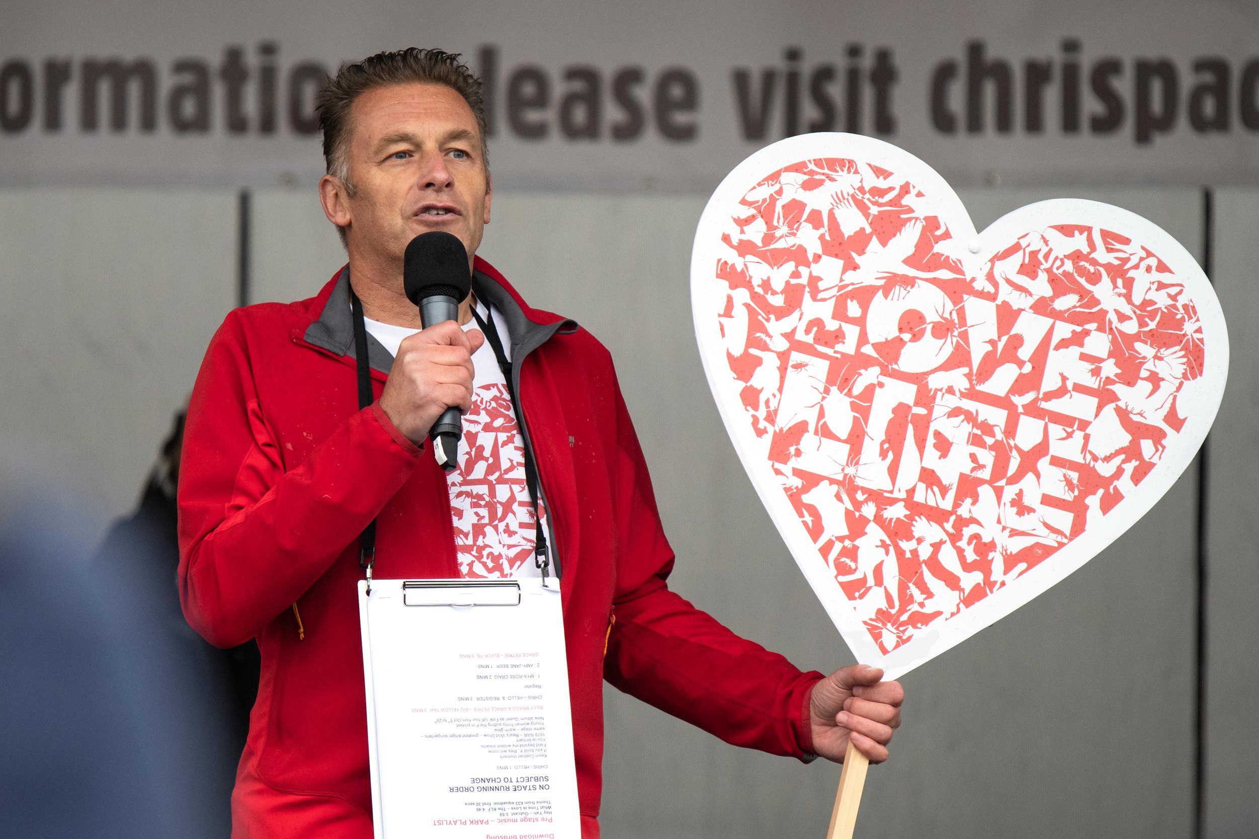 Chris Packham is speaking into a microphone and holding a heart-shaped sign that reads 'Love Life' with graphics of animals on it