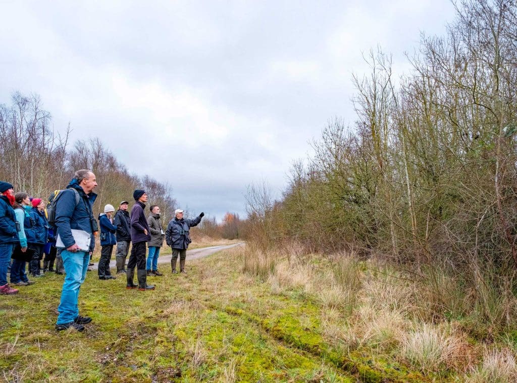A group of volunteers stood on a path looking into some bare trees whilst being trained by an instructor