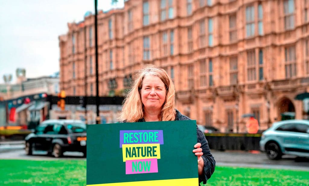A woman stands smiling at the camera holding a sign that reads 'Restore Nature Now'