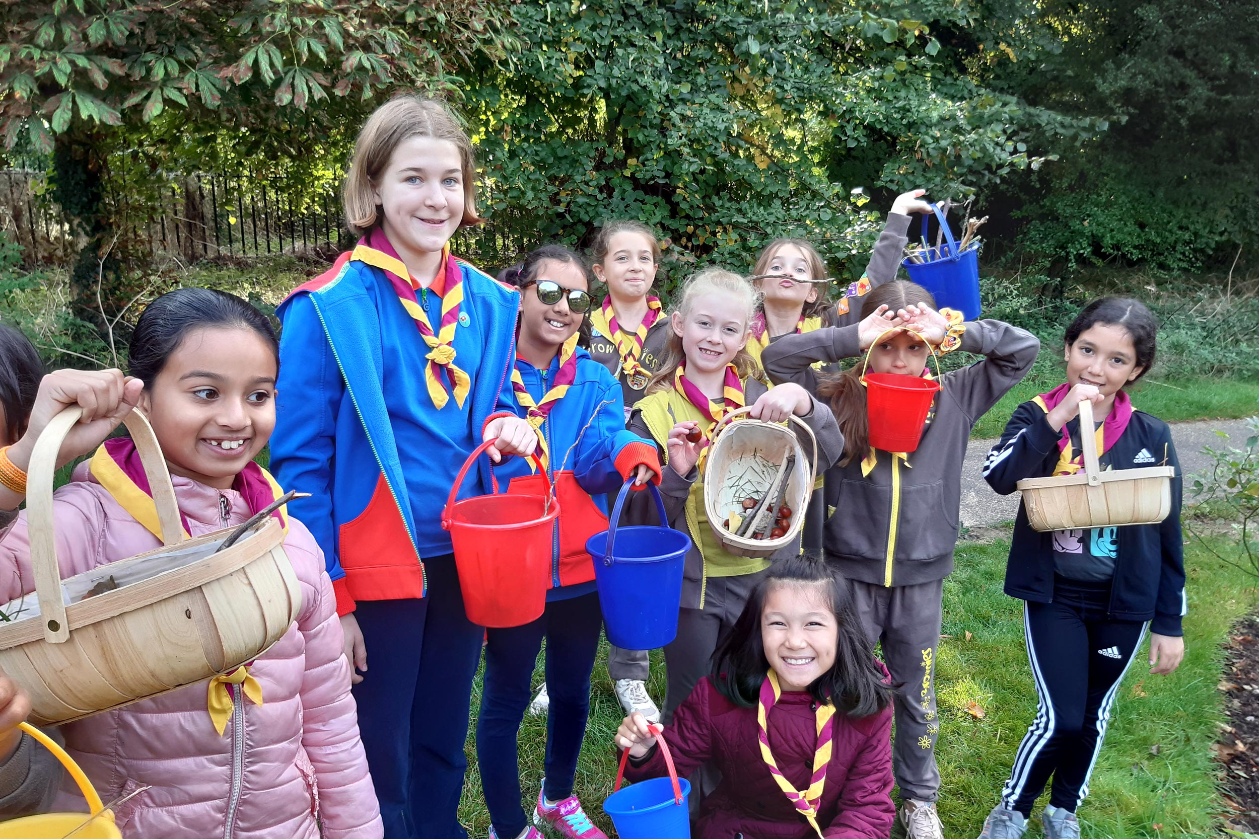 A group of young girls in Brownie and Girl Guide uniform stood in a park holding baskets and buckets of nature finds