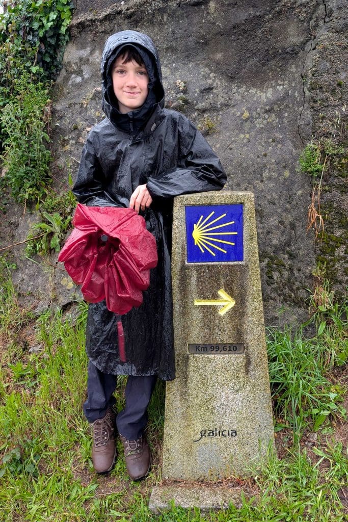 A young boy is stood leaning against a pillar on the Camino Ingles walking trail. He is wearing a hooded coat in the rain and holding an umbrella