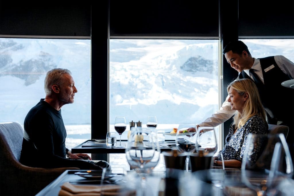 A couple being served in a cruise ship restaurant, snowy mountains in the background