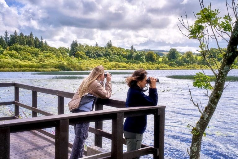 Two women stand on a boardwalk looking out over Lochwinnoch with binoculars