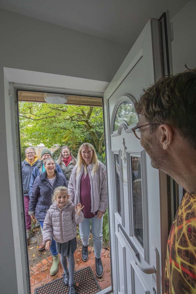 A man stands at an open front door welcoming in a group of friends