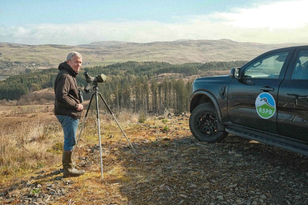 A man stands with a telescope overlooking a hilly landscape. He is smiling back towards the camera and a van next to him has an RSPB logo on it