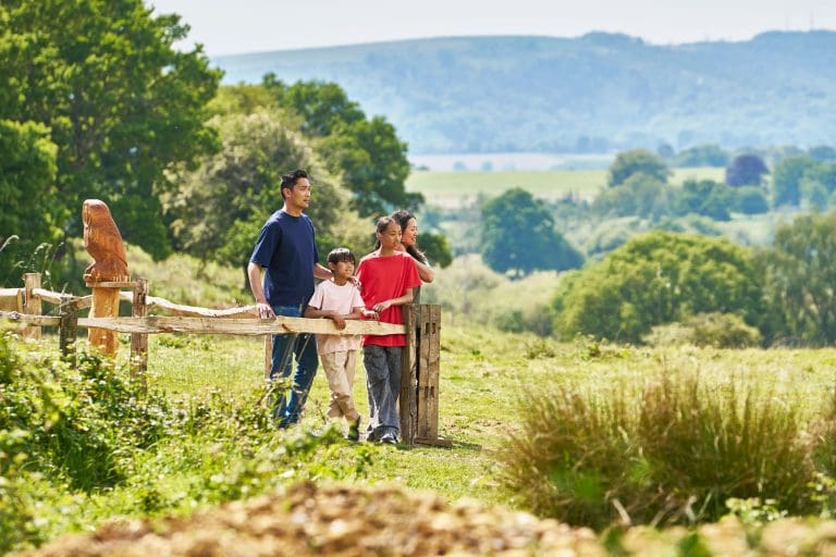 A family of four stand behind a fence in a large field looking into the distance. There are hills and trees in the background