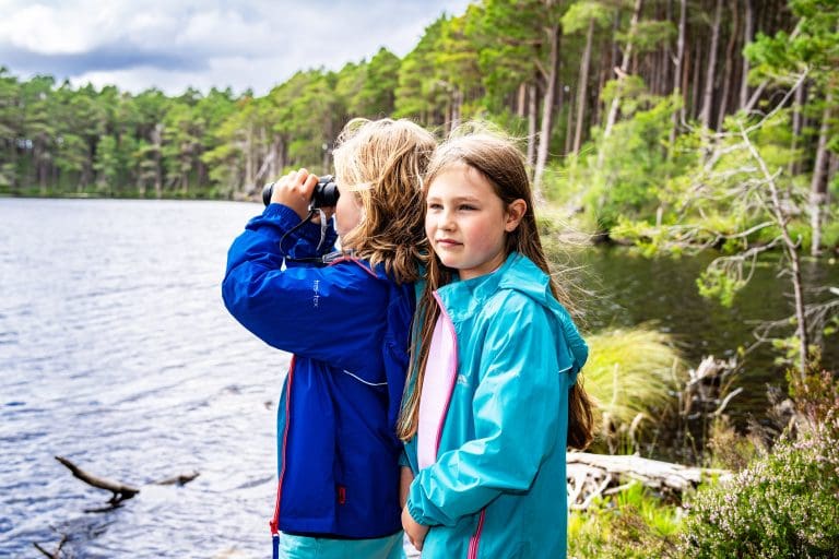 Two childrn stood next to a loch. One is looking out over the water through some binoculars