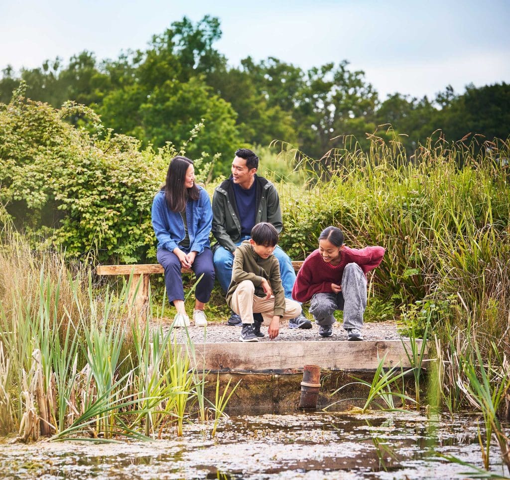 A family of four sat by a pond at a nature reserve. Two adults are sat on a bench and the children are crouched by the water smiling