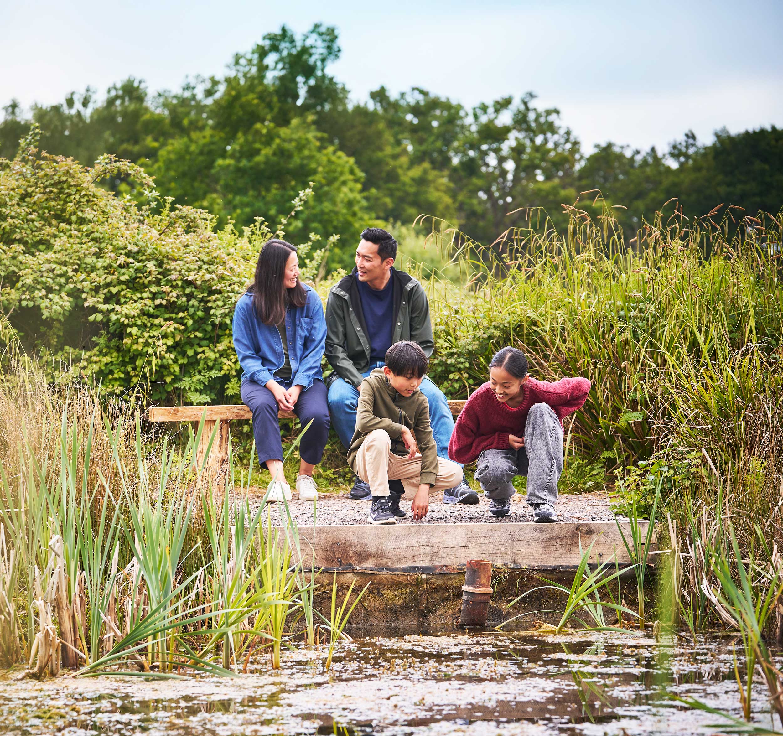 A family of four sat by a pond at a nature reserve. Two adults are sat on a bench and the children are crouched by the water smiling