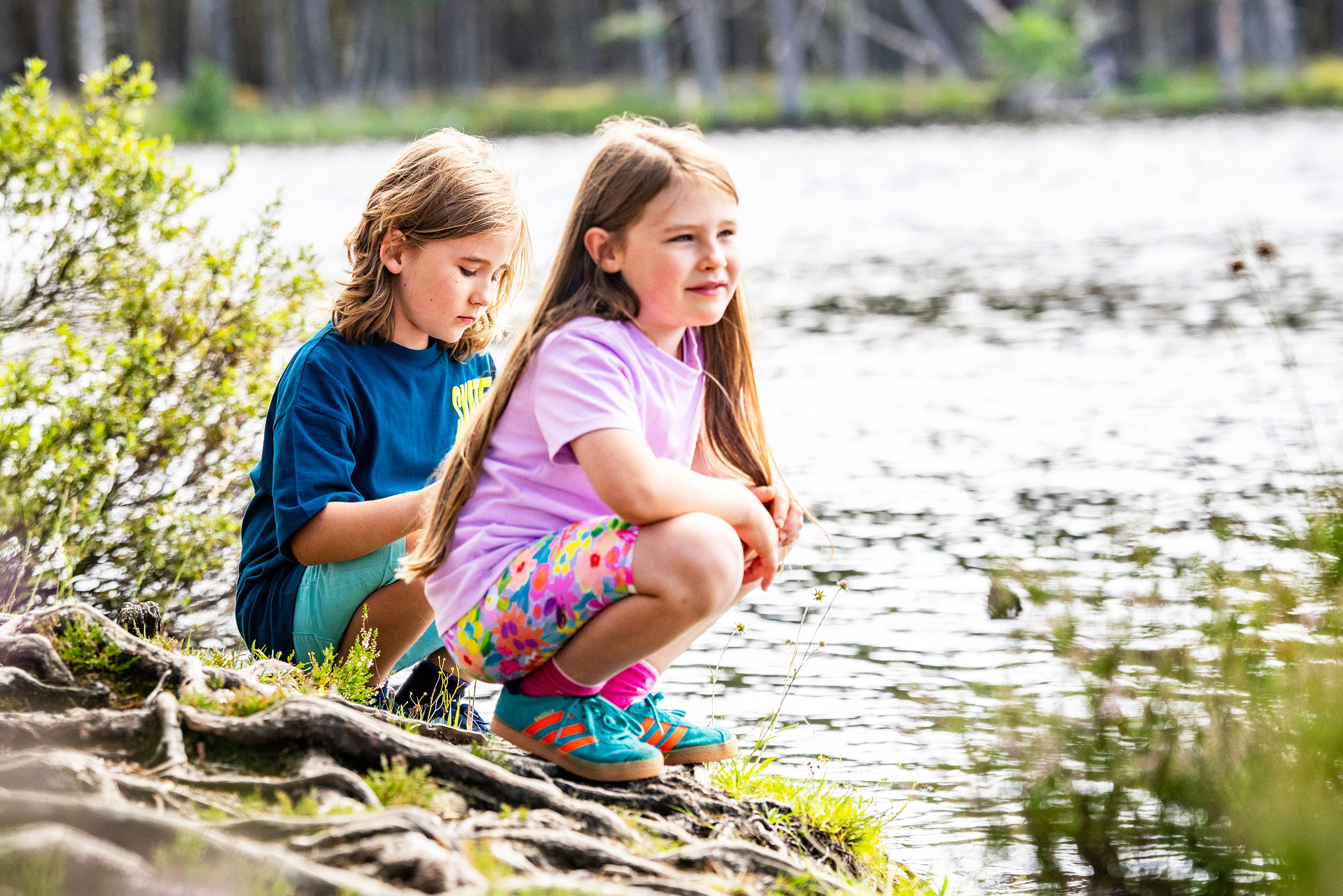 Two children crouch by a loch looking out over the water