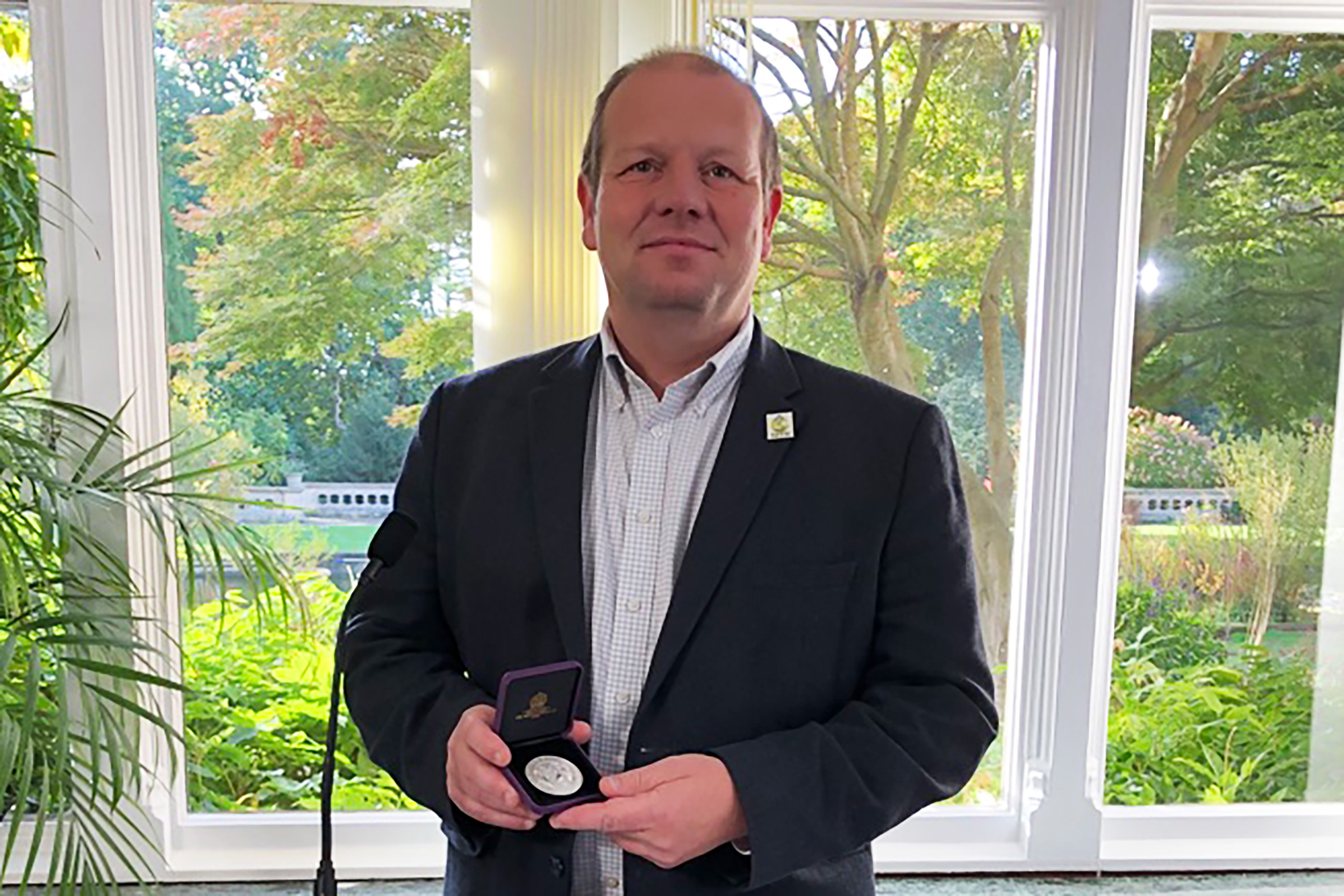 A man stands smiling in front of a window holding a silver medal presented in a case