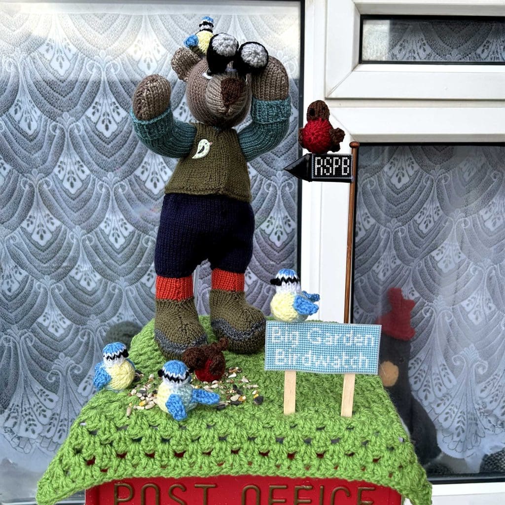 A knitted bear on top of a postbox. The bear is looking through binoculars and surrounded by knitted birds and a cross stitch 'Big Garden Birdwatch' sign