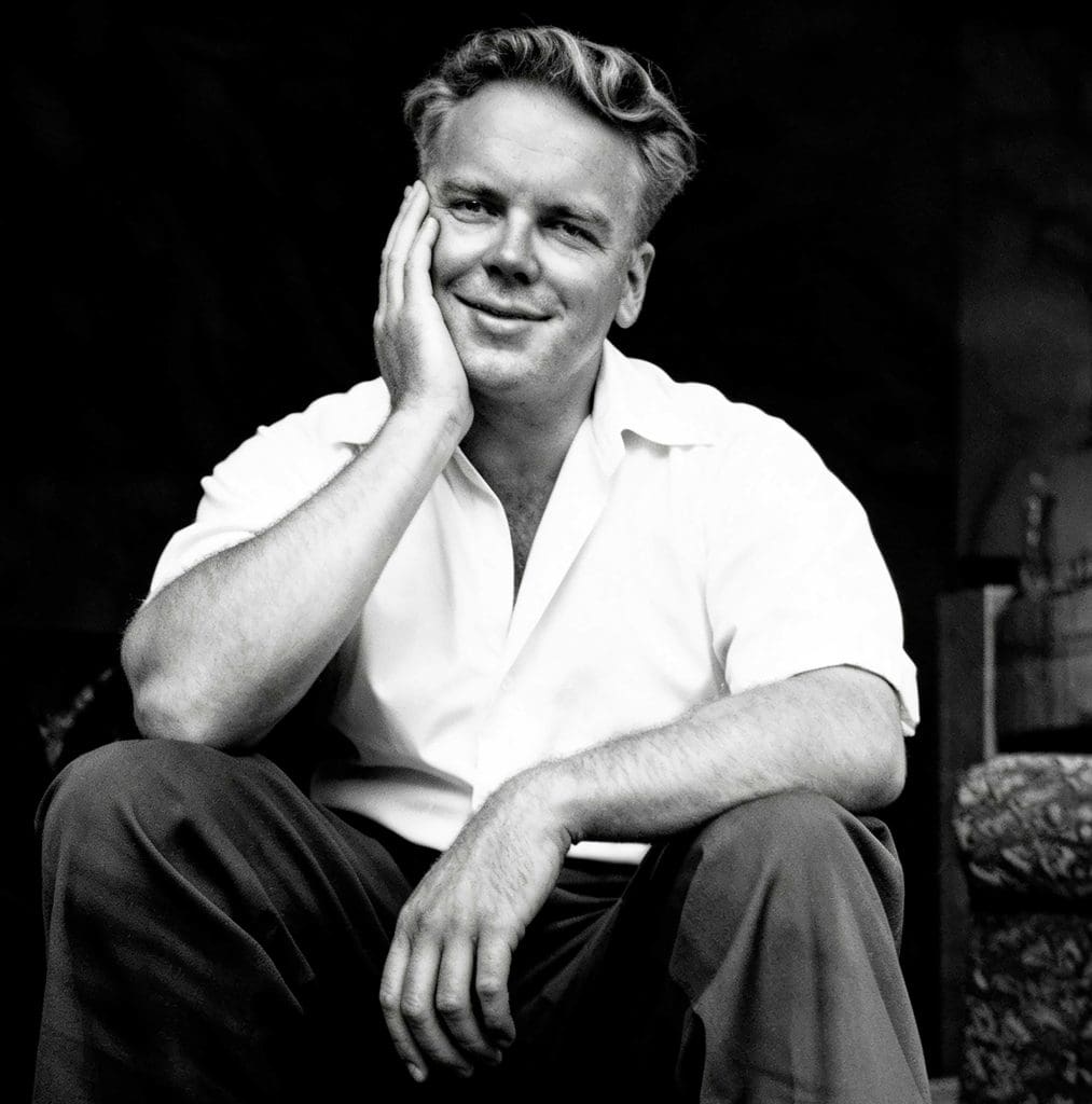 A black and white photo of author John Alec Baker, smiling, with his face resting on one hand, sitting in a living room