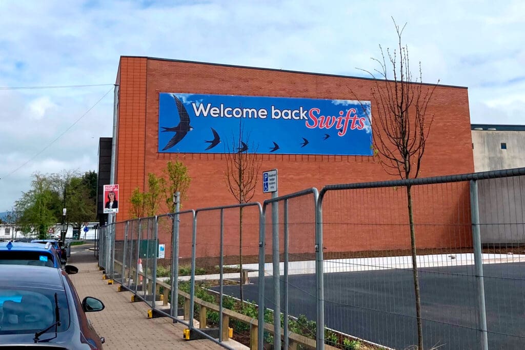 A huge banner reading 'Welcome back Swifts' on the side of a red brick building.