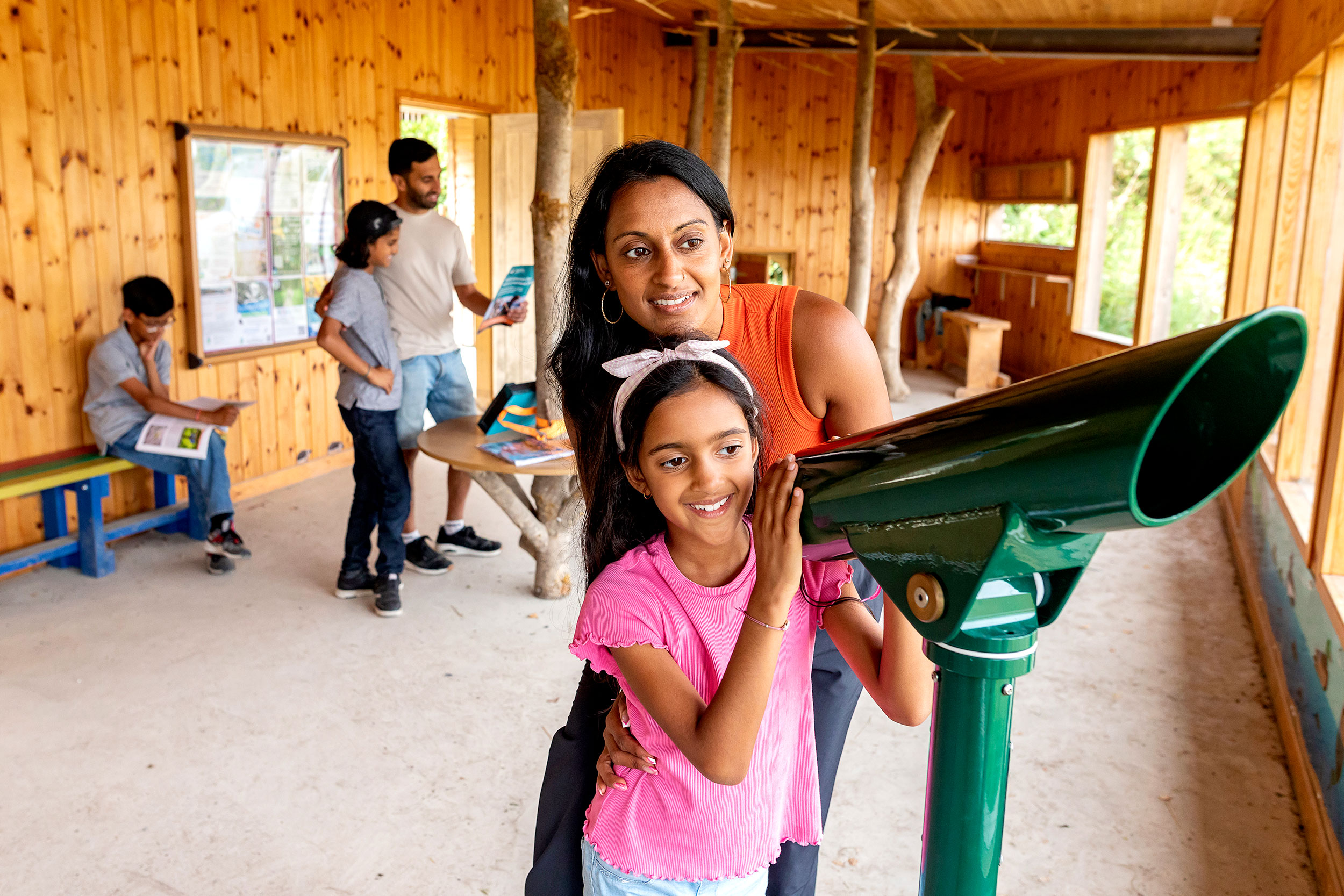 A woman and young girl stand in a bird hide next to a telescope, looking outwards