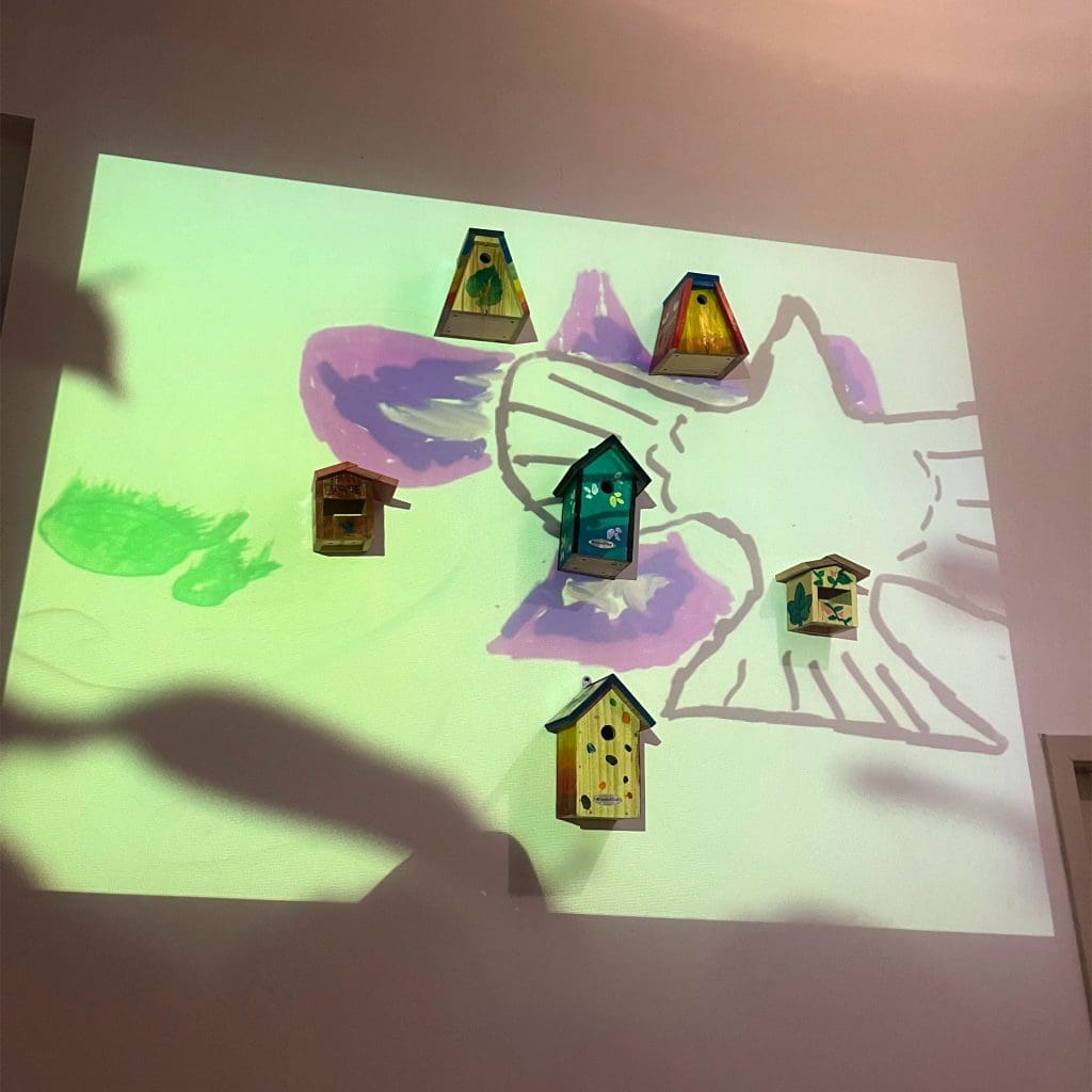 A piece of bird-inspired art: six bird boxes attached to a wall, arranged in a pentagon, with a painting of a bird projected over them