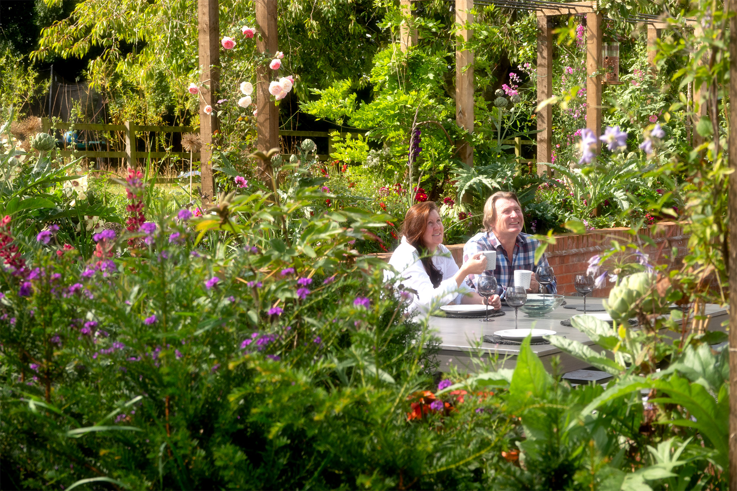 A woman and man, holding mugs, sitting at a table in their garden, surrounded by plants