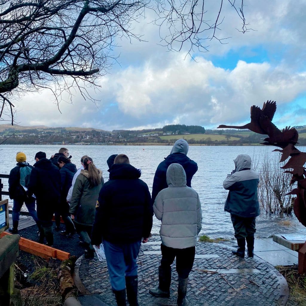 A group of around 10 young people walking onto a jetty next to a lake, on a cold day