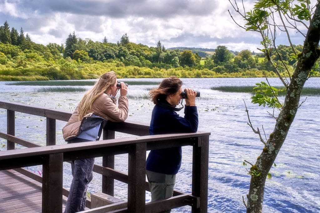 Two women stand on a viewpoint with binoculars overlooking a body of water.