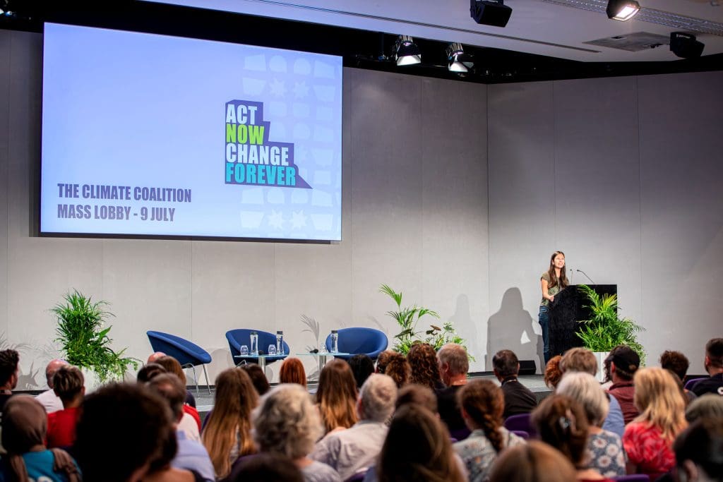 Mya-Rose Craig speaks to an audience at the mass lobby event. The powerpoint screen reads 'Act Now, Change Forever'