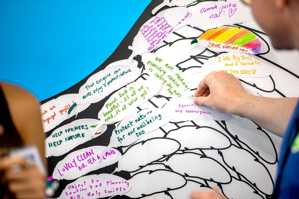 A craftivism project where people have written their hopes for the future of nature on feather shapes and compiled them into a big wing-shaped poster. The photo shows a hand sticking a feather onto the wings