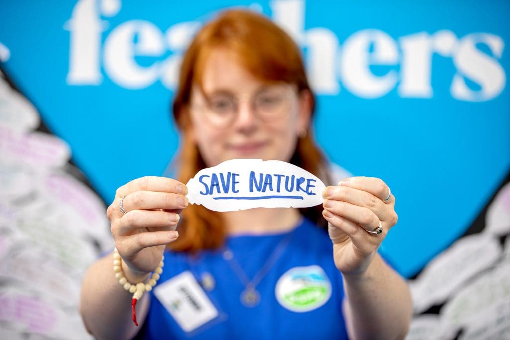 A young girl holds a paper feather that reads 'save nature'. She is out of focus in the background