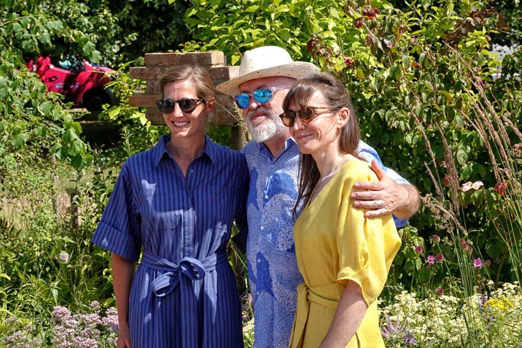 Comedian Bill Bailey stood in the RHS Swift Garden with his arms around two women. All three are smiling at a camera