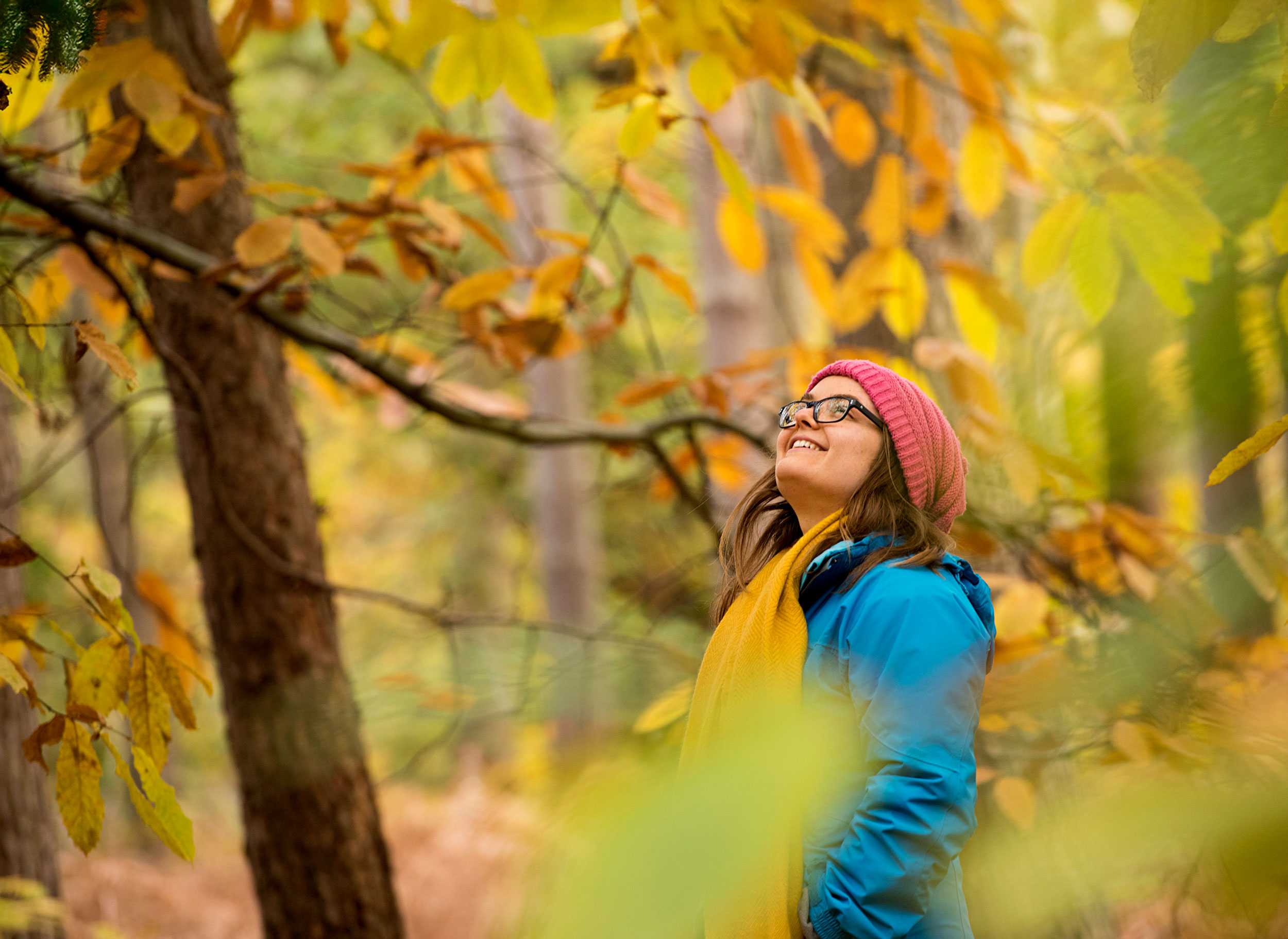 A women wearing a coat, hat and scarf is stood amongst trees with autumnal leave. She is looking up and smiling.