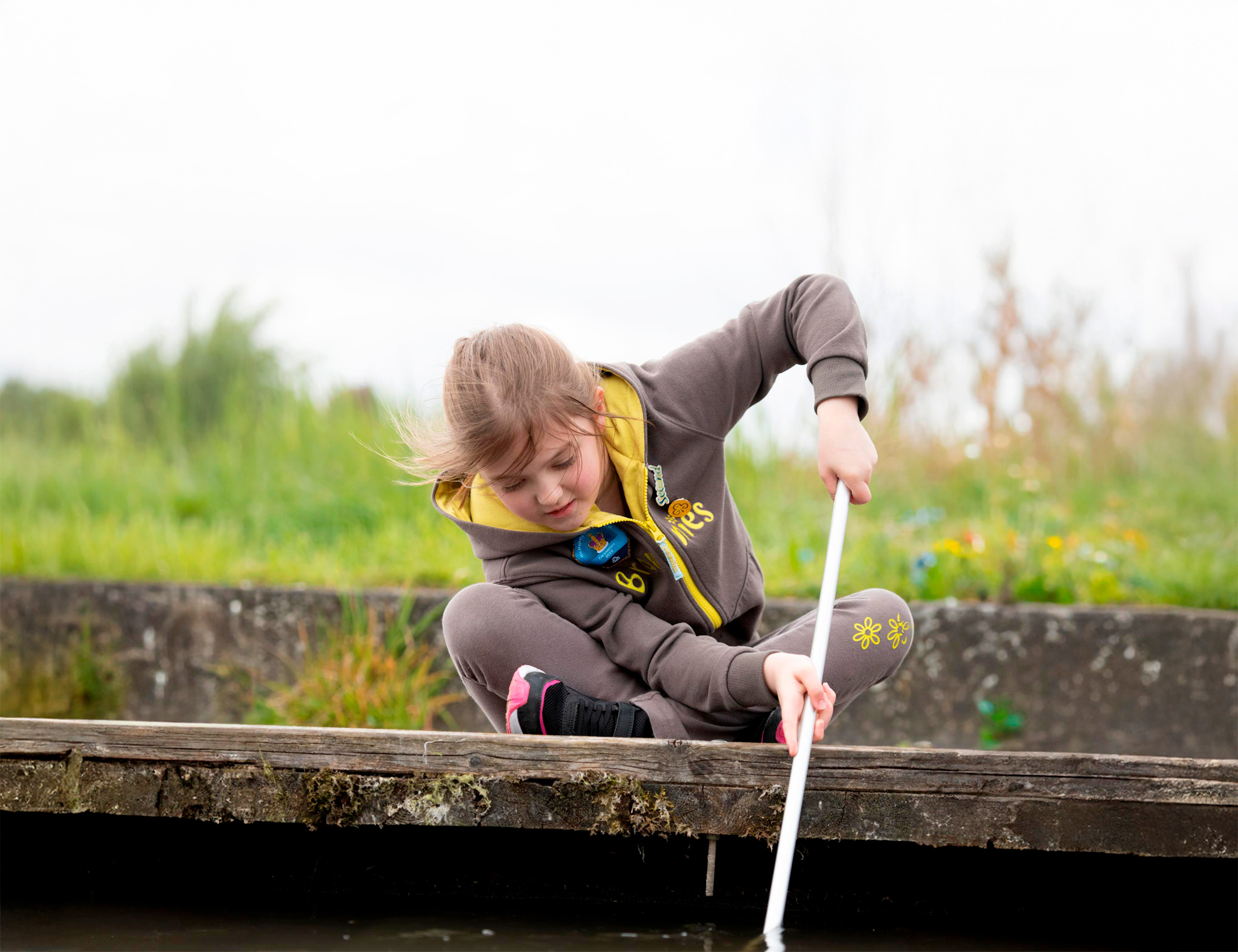 A young girl in a Brownies uniform is sat on a boardwalk dipping a net into water